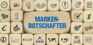 Viele Holzwuerfel mit Symbolen aus dem Marketing In der Mitte steht in blauer Schrift "Markenbotschafter"