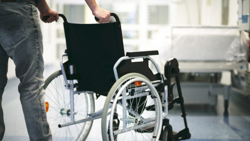 Zu sehen ist ein leerer Rollstuhl der von einer mänlichen Person in ein Zimmer mit Krankenbett geschoben wird.