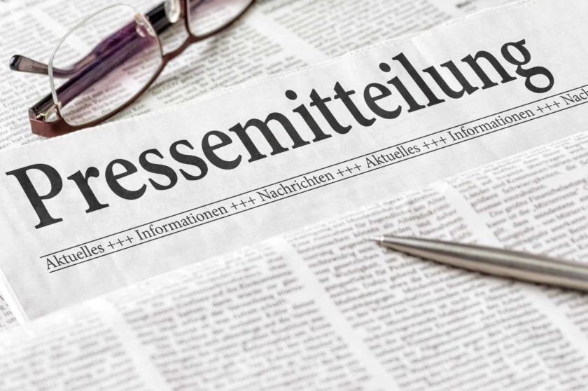 Das Bild zeigt eine Zeitung, ein Teil einer Brille und einen Kugelschreiber. Auf der Zeitung steht in großer Überschrift " Pressemitteilung".