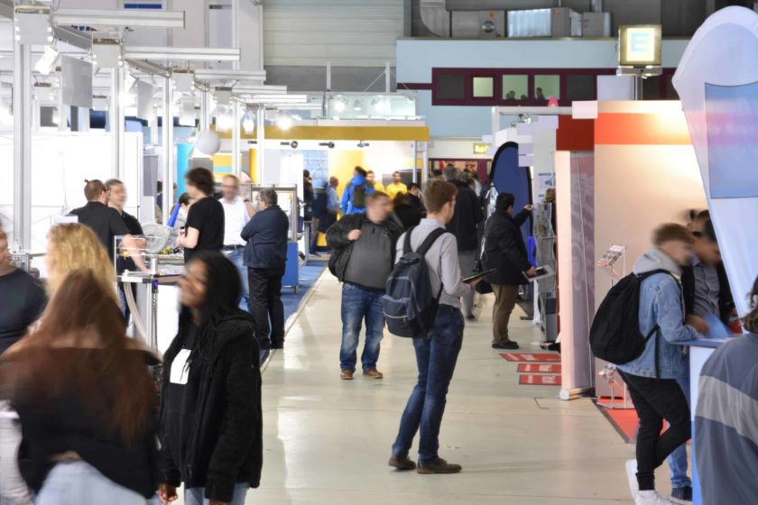 Das Bild zeigt den Blick in eine Fachmesse mit Personen an Ständen.