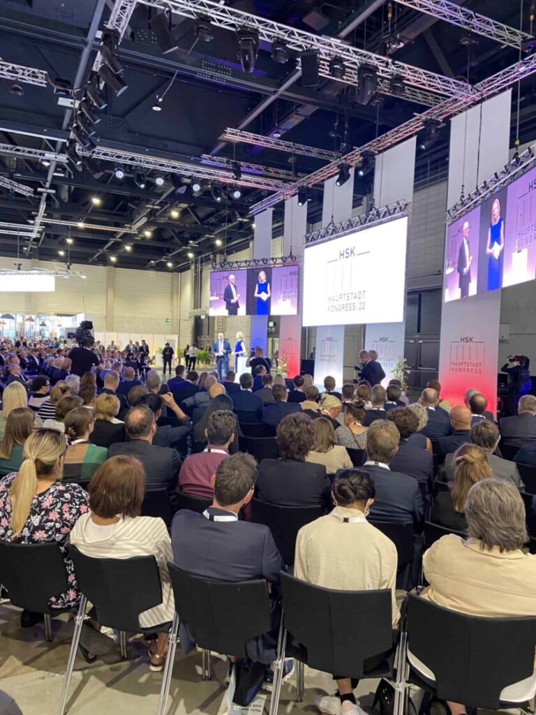 Das Bild zeigt die Eröffnungsveranstaltung Hauptstadtkongress 2022. Zahlreiche Personen sitzen mit der Blickrichtung zum Podium in einer Halle.